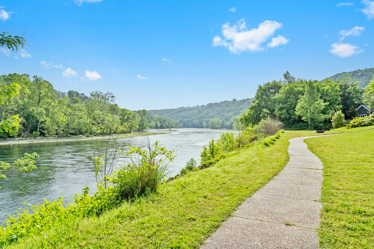 A peaceful riverside pathway meanders through trees and gentle hills, suggesting opportunities for scenic walks, fishing, or peaceful reflection while enjoying the outdoors. The natural setting is calm and welcoming.