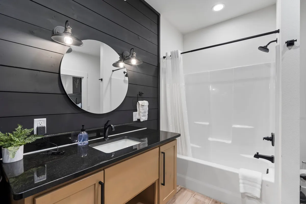 A modern private bathroom with a black shiplap accent wall, circular mirror, stylish sink, and elegant light fixtures, creating a calming and refreshing ambiance.