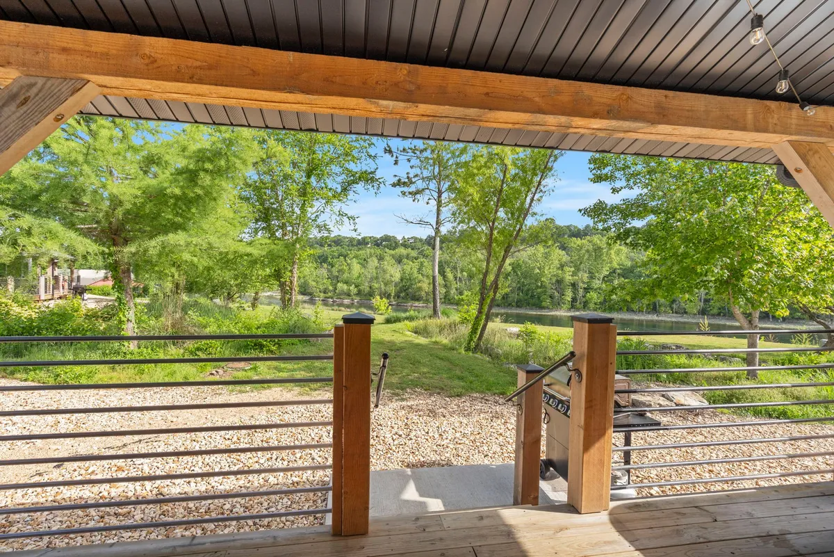 An inviting exterior scene with a wooden railing and gravel path leading through lush greenery toward a sparkling lake view. The space is perfect for relaxing walks, wildlife watching, or casual outdoor dining.