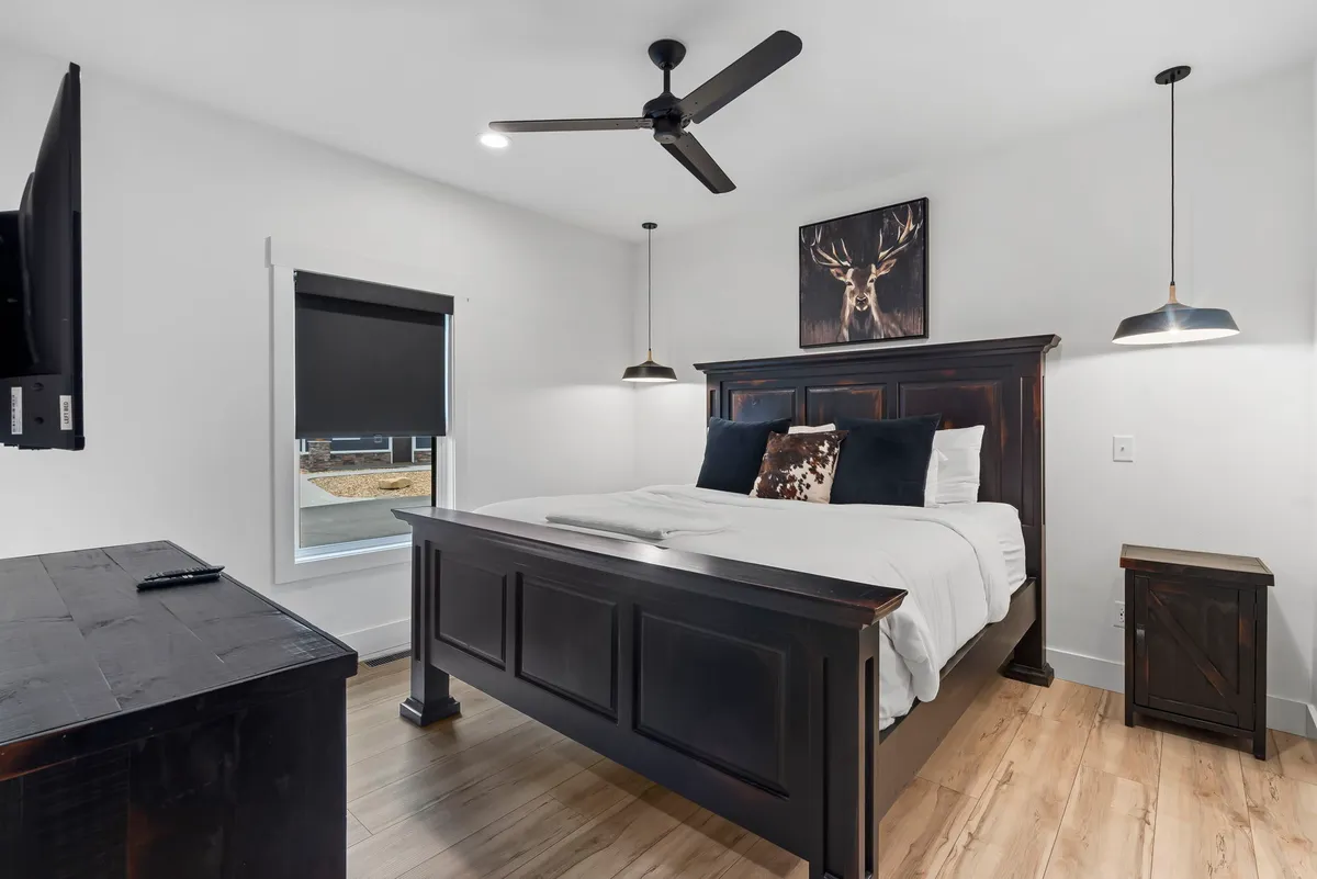 A serene king bedroom featuring a plush bed with cozy linens, deer artwork above the headboard, pendant lighting, and a wall-mounted television. The space exudes comfort and relaxation, perfect for restful nights.