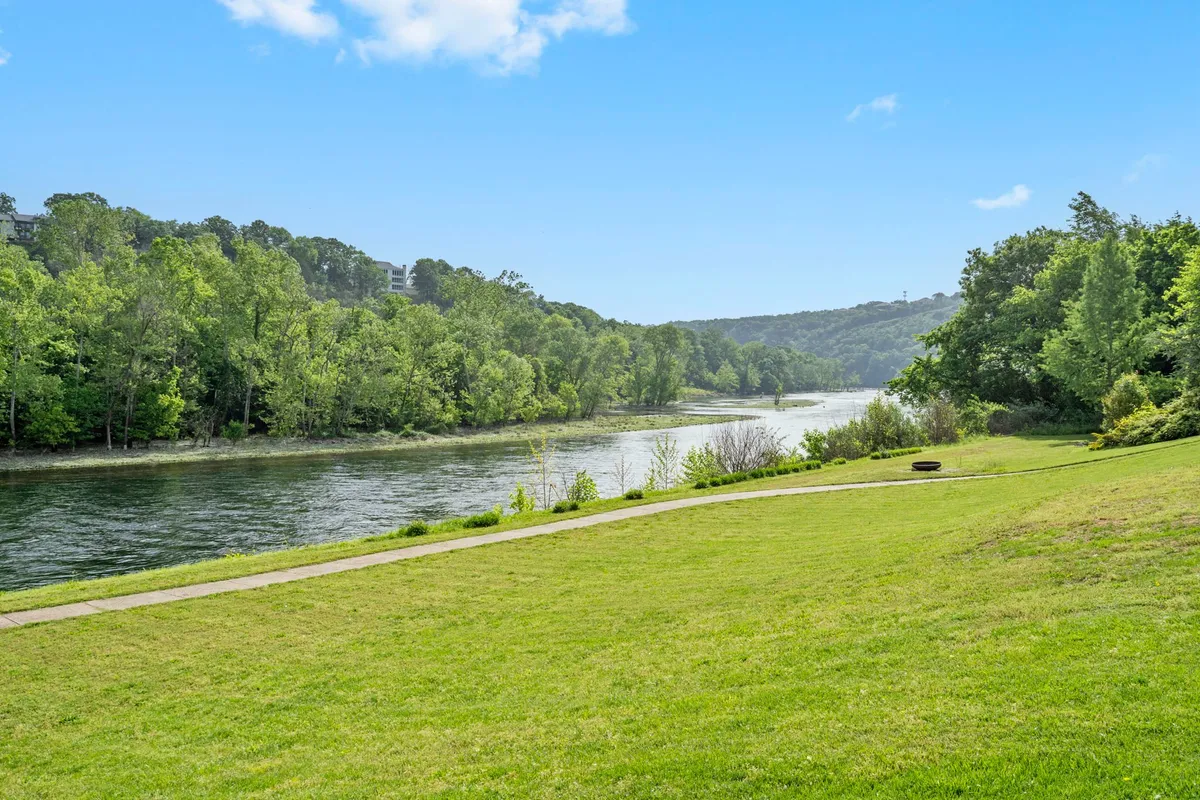 A broad, serene view of the river bordered by lush green trees and an open lawn, inviting families to picnic, fish, or simply relax. The scene hints at space for outdoor games and gatherings, with the water easily accessible.