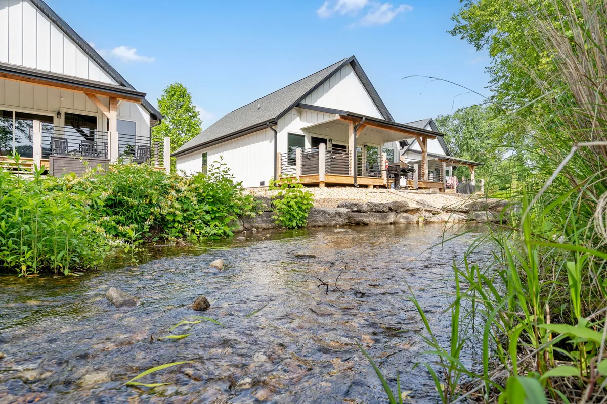 This image captures the porches of several houses set alongside a lush stream, with people relaxing by the water or enjoying meals on their porch. The natural greenery and waterway create a peaceful, rejuvenating mood.