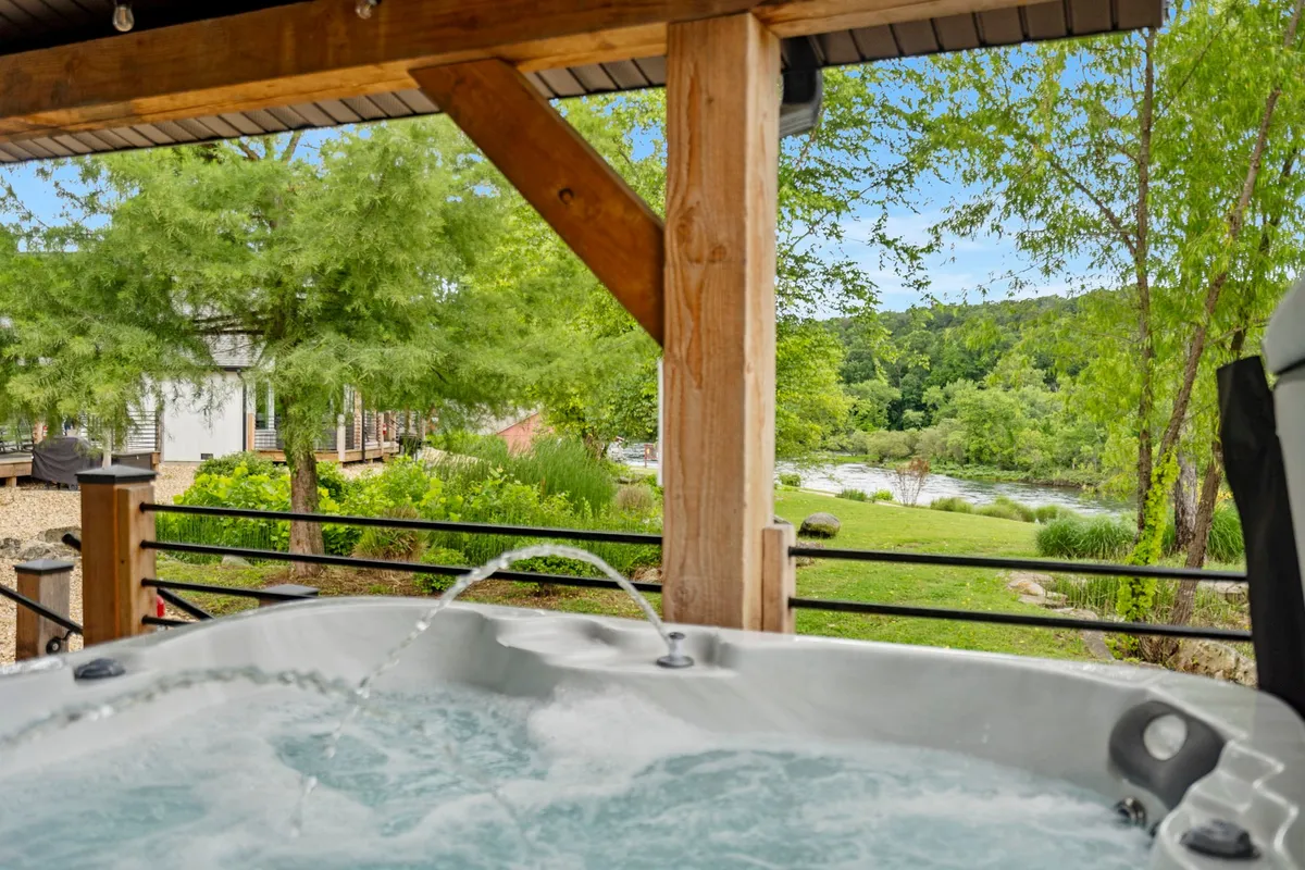 This exterior scene captures a private hot tub set amidst lush greenery, overlooking a serene river. The tranquil environment invites guests to unwind while soaking in stunning natural views at sunset or under the stars.