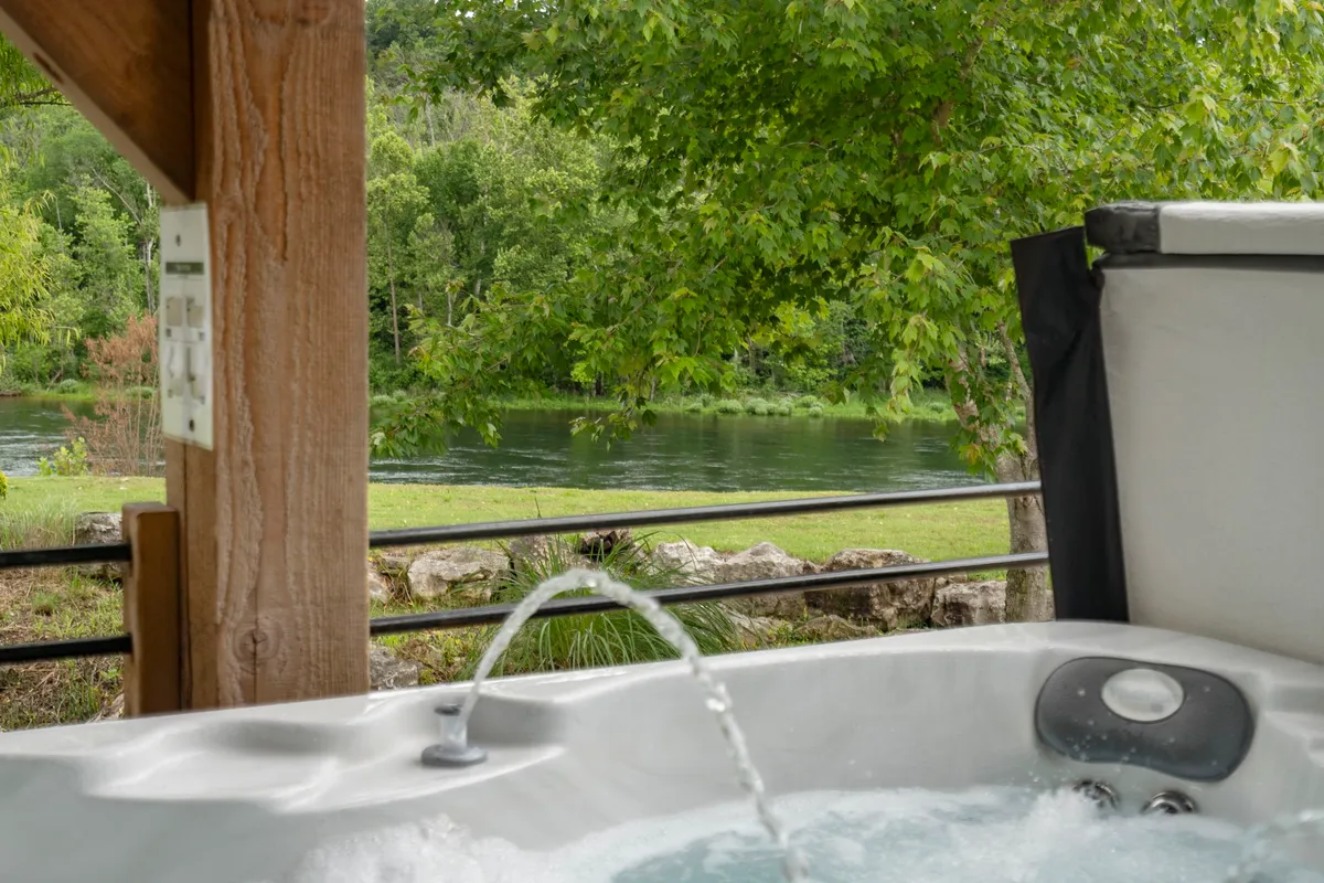 This image captures a private hot tub set on a patio with lush greenery and a river view in the background. The setting is serene, with the hot tub being the focal point and inviting guests to relax while soaking in the natural beauty. The patio appears spacious, offering a perfect spot for quiet moments or socializing.