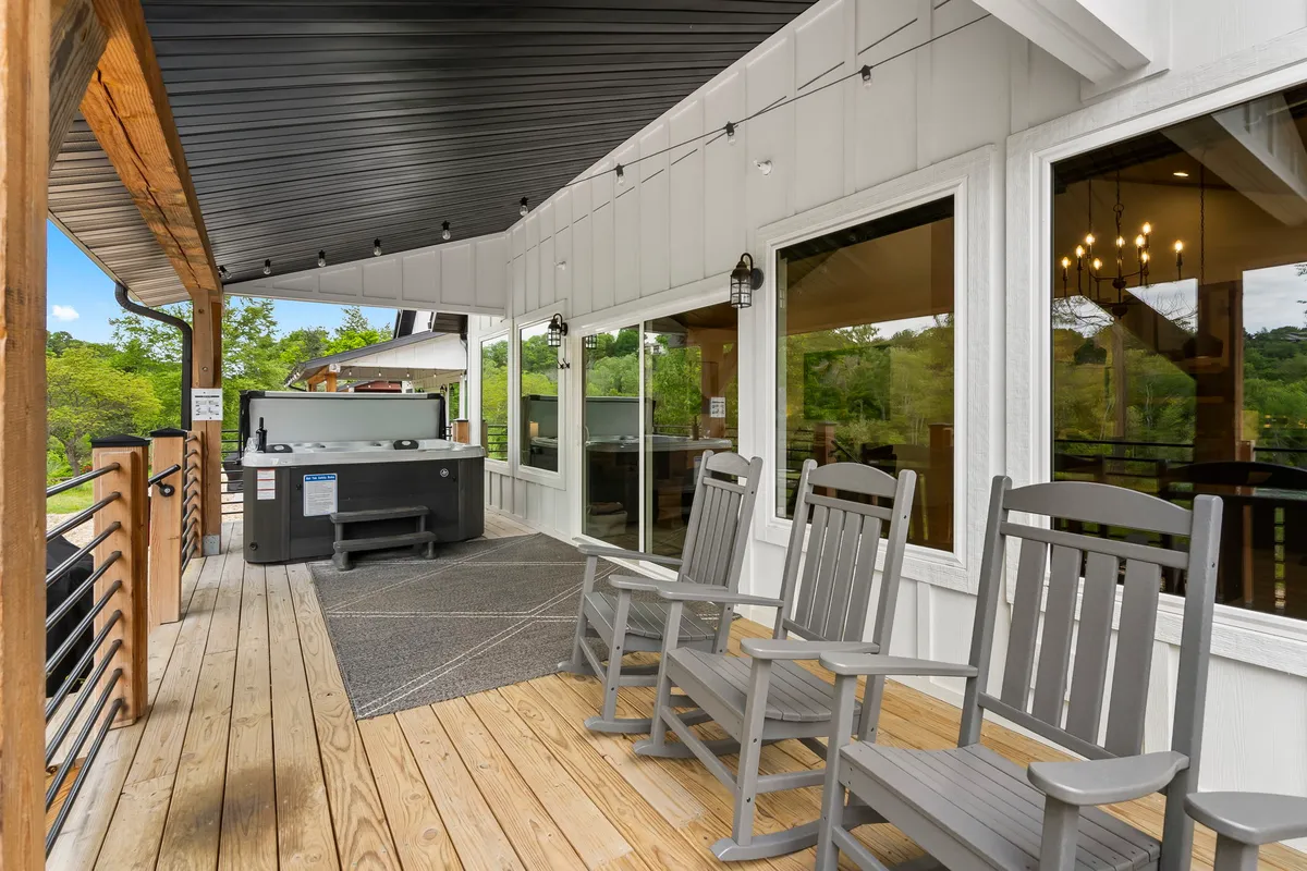 This image shows a spacious deck with a hot tub, rocking chairs, large windows, and a deck railing. The scene is inviting, with hints of sunset lighting, and the arrangement highlights both relaxation in the hot tub and socializing in the seating area.