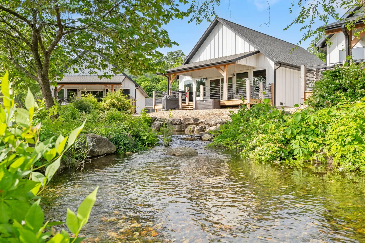 The image showcases a modern lakefront house nestled amid lush landscaping, with a peaceful stream running alongside the property. The scene exudes serenity, with the inviting exterior of the home and natural waterfront setting suggesting relaxation and outdoor enjoyment.
