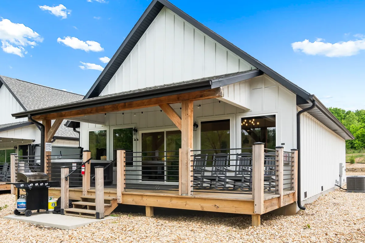 The photo features a covered porch with comfortable rocking chairs and an outdoor grill, overlooking a green natural setting. The grill is prominently placed, emphasizing outdoor cooking and dining opportunities, while the cozy seating invites guests to enjoy the Ozarks breeze and nature sounds.